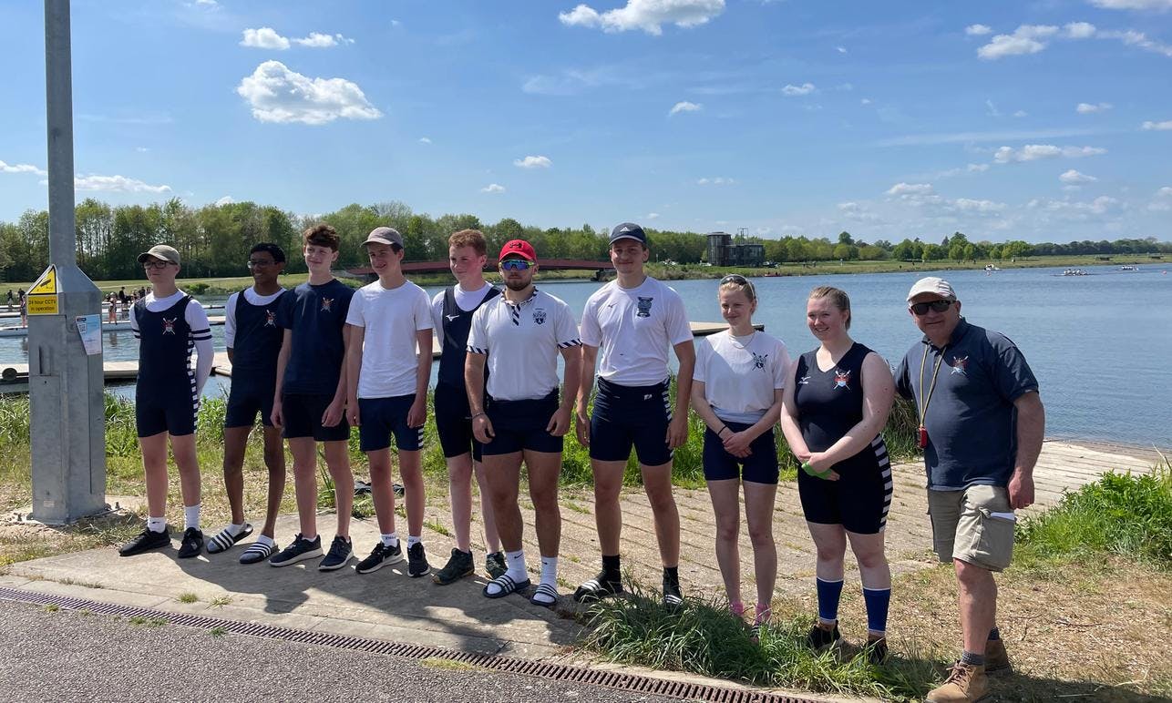 Nine juniors and a coach standing in the sunshine in front of the vast rowing lake at Dorney. 
